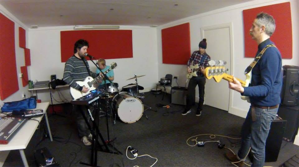 Four of the band rehearsing in a room lined with red acoustic panels, two guitars, a bass and a Novation keyboard between them