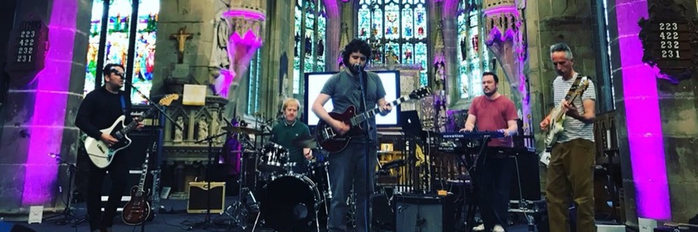 Five of the band playing in a church, stone pillars lit purple and stained glass behind them