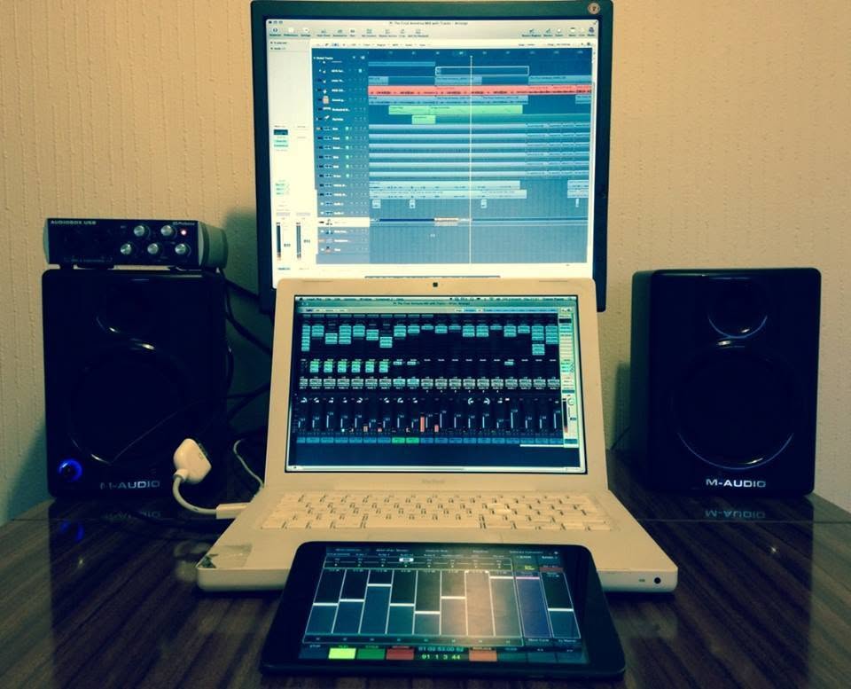 A white MacBook and a second screen on a desk between M-Audio monitors, a session open across both and an iPad running a mixer in front