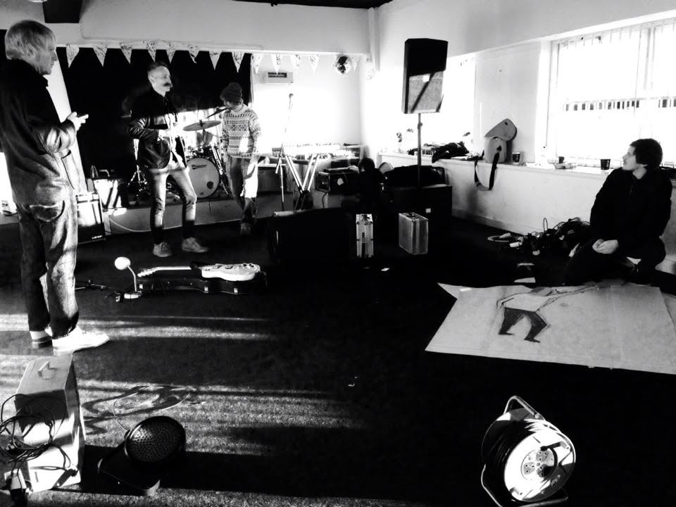 Black and white photograph of the band setting up for the album launch in a bare room hung with bunting, flight cases and a hand-drawn backdrop laid out on the floor